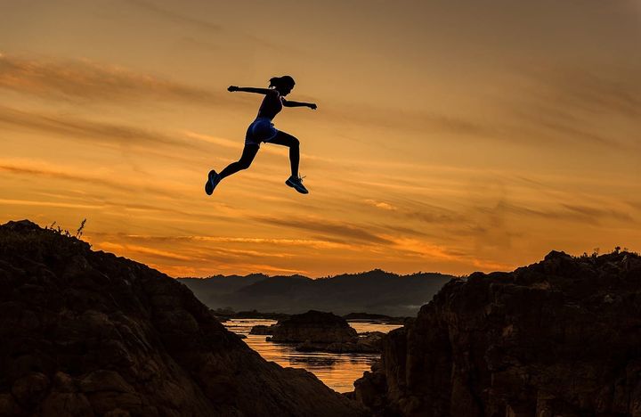Silhouette d’une femme qui saute d’un rocher à l’autre au coucher du soleil, au-dessus d’une rivière