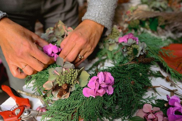 Mains qui composent une couronne avec des branches de conifère et des hortensias roses, un sécateur orange au premier plan